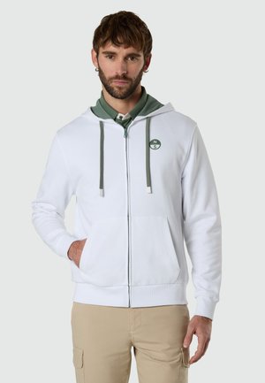 Uomo con barba che indossa una felpa con cappuccio bianca con zip sopra una maglietta verde, in piedi con una mano in tasca contro uno sfondo neutro.