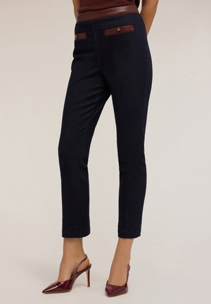 Pantaloni attillati blu navy con una texture liscia, dettagli in pelle marrone sulle tasche e accessori in tonalità oro. Abbinati a décolleté borgogna.