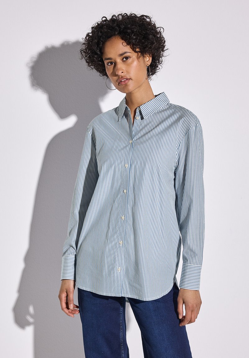 Camisa de botones a rayas azules y blancas con mangas largas, cuello y bolsillo; combinada con jeans azul oscuro. Textura suave, corte regular.