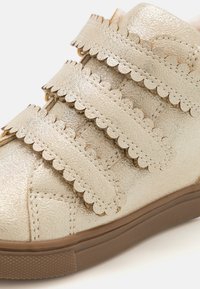 Guldmetalliska barnsneakers med tre vågiga kardborreband, texturerad ovandel och en brun gummisula.