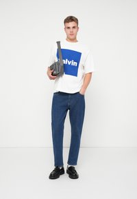 Młody mężczyzna w białym T-shircie z niebieskim nadrukiem, niebieskich dżinsach, czarnych mokasynach, trzymający szaro-skorzaną torbę na ramię na białym tle.