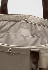 Bolso tote de color beige claro con un interior liso y texturizado. Presenta el logo de Lululemon y un cierre de presión, acentuado por correas de tela marrón oscuro.