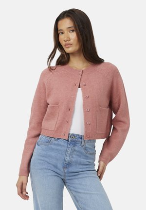 Femme portant un cardigan rose boutonné avec poches avant sur un haut blanc et un jean taille haute bleu clair, debout devant un fond blanc.