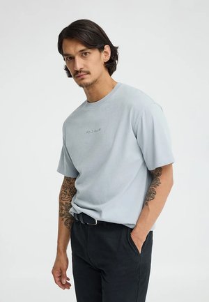 Mężczyzna o ciemnych włosach i tatuażach na ramionach, ubrany w jasnoniebieską koszulkę "Polo Club" i czarne spodnie, stoi z jedną ręką w kieszeni.