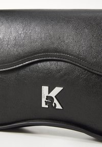 Juodas dirbtinės odos delninukas su tekstūruota paviršiumi, kuriame yra sidabrinis iškilus 'K' logotipas ir bangos dizaino detalė.