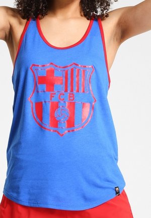 Osoba nosząca niebieski tank top z czerwonym obszyciem i dużym czerwonym logo FC Barcelona, połączonym z czerwonymi szortami.