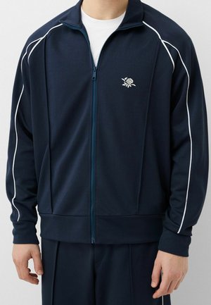 Mann trägt eine marineblaue Trainingsjacke mit Reißverschluss, weißer Paspelierung und einem kleinen weißen Rosenemblem auf der Brust, kombiniert mit passenden Hosen.