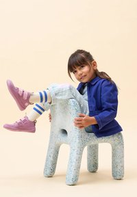 Kind sitzt auf einem gesprenkelten blau-grünen Plastikhocker, trägt eine blaue Jacke, pastellfarbene Hosen, lila Schuhe und weiße Socken mit blauen Streifen.