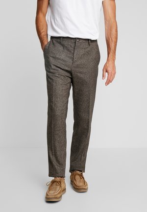 Mand iført brune teksturerede bukser, tanfarvede snørestøvler og en hvid indstikket skjorte, stående med den ene hånd i lommen.