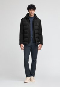 Svart pufferjacka med huva, med dragkedjeöppning och sydda detaljer. Bärs över en mörkblå tröja och matchas med mörka byxor.