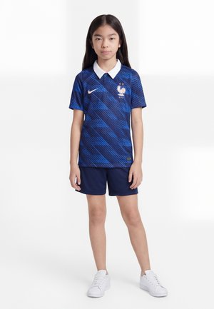 Chica de pie de frente, lleva una camiseta de fútbol de Francia azul con estampado y cuello blanco, pantalones cortos azules a juego y zapatillas blancas sobre fondo blanco.