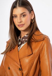 Abrigo de cuero marrón con doble botonadura y amplios solapas, con botones metálicos. Debajo, se asoma una blusa con estampado y un cuello oscuro.