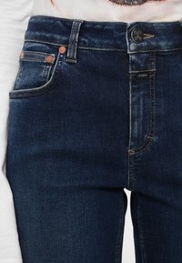 Gros plan sur un jean en denim bleu foncé avec rivets en cuivre, poche avant et bouton étiqueté "CLOSED."