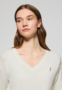 Pull crème à col en V avec une coupe ample, un col côtelé et un petit logo brodé sur le côté gauche. Le tissu semble doux et lisse.