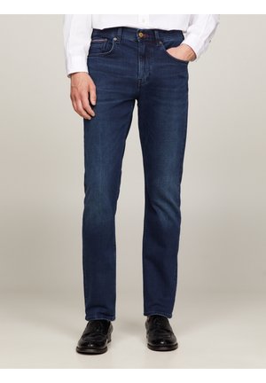 Homme portant un jean droit bleu foncé, une chemise blanche et des chaussures habillées noires, debout avec une main dans la poche devant un fond neutre.
