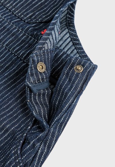 Salopettes en denim bleu marine foncé avec des rayures blanches. Présente des accessoires en métal et des accents de couture rouge. La texture du tissu semble robuste et tissée.