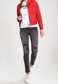 Červená bomber bunda, bílý sweatshirt s grafikou, roztrhané černé upnuté džíny a bílé tenisky. Texturovaný materiál, přiléhavý střih, s viditelným poškozením.