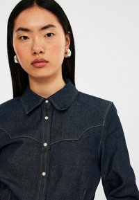Mørk denimskjorte med knaplukning foran, spidse kraver og kontrasterende syninger. Har et yolk-design og sølvhardware.
