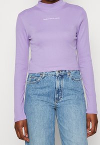 Osoba nosící těsně přiléhající levandulový crop top s dlouhými rukávy a nápisem „MARC O'POLO DENIM“ a vysokým pasem a světle modré džíny.