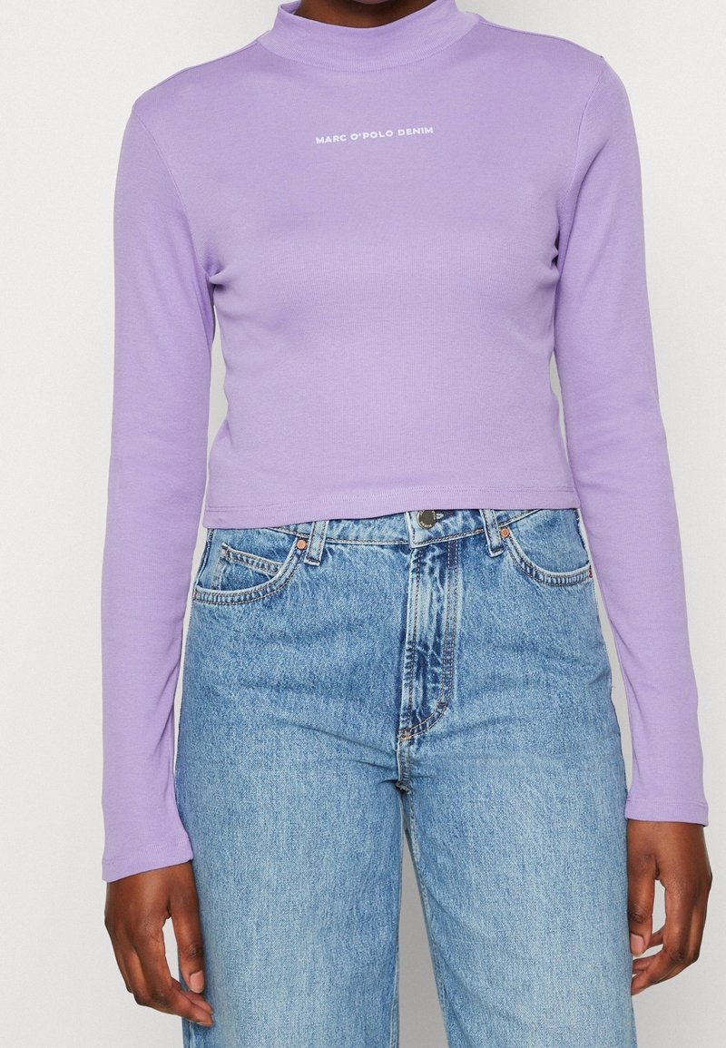 Osoba nosící těsně přiléhající levandulový crop top s dlouhými rukávy a nápisem „MARC O'POLO DENIM“ a vysokým pasem a světle modré džíny.