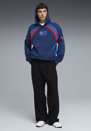 Joven con sudadera azul con rayas rojas y azul claro, pantalones negros de pierna ancha y zapatillas blancas, de pie con las manos en los bolsillos.