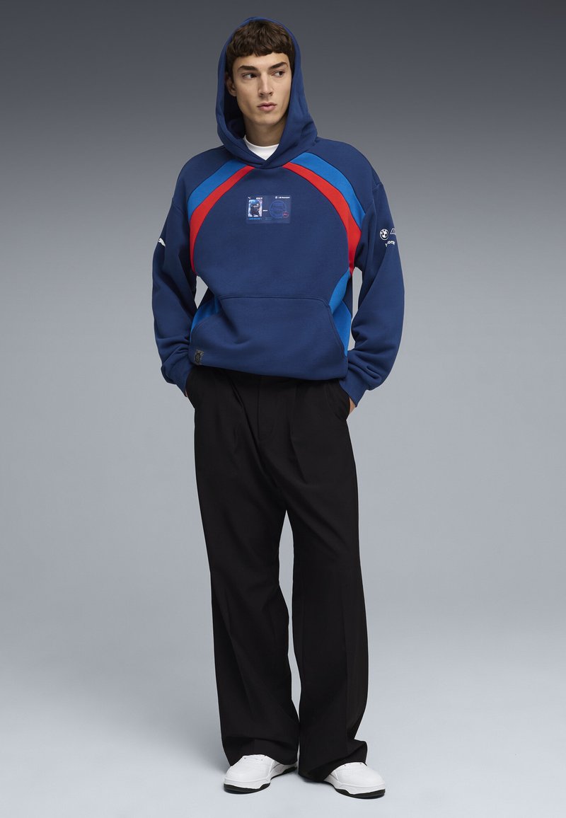 Nuori mies, jolla on sininen huppari, jossa on punaisia ja vaaleansinisiä raitoja, mustat leveälahkeiset housut ja valkoiset lenkkarit, seisoo kädet taskuissa.