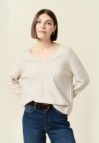 Vrouw met een steile bruine bob-kapsel, die gouden oorbellen draagt, een beige V-hals trui en een blauwe spijkerbroek met een patroongordel.