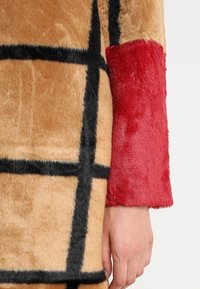 Prenda de piel sintética con base beige y patrón de cuadrícula negra, y una manga texturizada de color rojo brillante. Superficie suave y táctil con colores intensos.