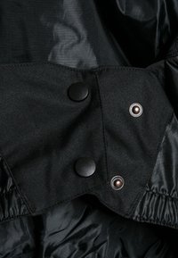 Wnętrze czarnej kurtki wykonane z gładkiej tkaniny, wyposażone w dwa czarne zatrzaski oraz brązowy metaliczny zatrzask z ozdobnym brzegiem.