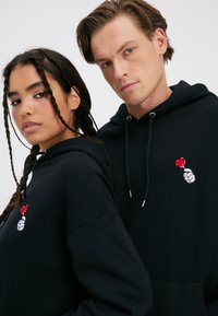 Black hoodies made of soft material feature a graphic of a hand holding a red heart, with silver eyelets and drawstrings.
