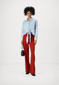 Ljusblå denimskjorta med knuten fram, matchad med högmidjade röda byxor. Modellen håller en liten svart axelväska och bär svarta skor.