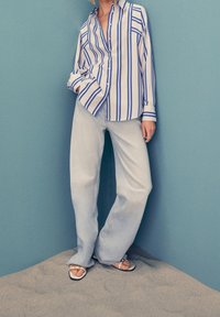 Persona che indossa jeans larghi di colore azzurro chiaro, sandali bianchi e una camicia bianca con righe verticali blu, in piedi sulla sabbia davanti a un muro blu.