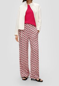 Weiße Zip-Jacke über einem pinken, gerippten Oberteil, kombiniert mit weiten gemusterten Hosen mit pinken und schwarzen Blattformen. Schwarze Absatzsandalen.