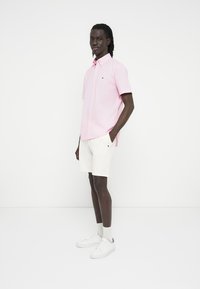 Ροζ κοντομάνικο πουκάμισο με κουμπιά και λογότυπο στο στήθος, σε συνδυασμό με λευκά σορτς και λευκά αθλητικά παπούτσια, στέκεται σε απλό φόντο.