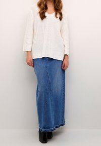 Femme portant un pull en tricot blanc, une longue jupe en denim bleu avec un ourlet effiloché, et des bottines noires, debout devant un fond uni.