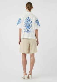 Hvit t-skjorte med korte ermer som har blå blomsterbroderi på fronten og ermene, parret med beige shorts og hvite sko med blokkhel.