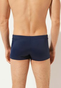 Marineblaue Badeshorts aus glattem Material, mit enger Passform und minimalen Nahtdetails. Die Rückansicht zeigt einen tief geschnittenen Bund.