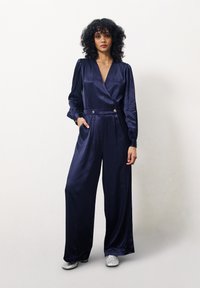 Combinaison en satin navy avec de longues manches bouffantes, décolleté en V profond et jambes larges plissées. Présente des boutons décoratifs et des poches latérales, associée à des chaussures étincelantes.