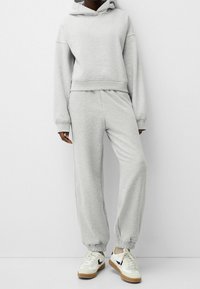 Sudadera gris claro con un bolsillo canguro y puños elásticos, combinada con pantalones deportivos gris a juego. Ambos hechos de una suave mezcla de algodón.