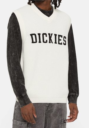 Homme noir portant un gilet en tricot blanc sans manches avec "DICKIES" en lettres noires sur une chemise noire à manches longues et un pantalon gris.