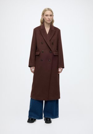 Femme portant un long manteau marron foncé à double boutonnage sur un jean bleu à jambes larges et des chaussures noires, debout devant un fond blanc uni.