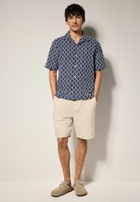Cămașă cu mânecă scurtă de culoare bleumarin cu un model geometric alb, asortată cu pantaloni scurți bej deschis și adidași gri slip-on. Design simplu și croială lejeră.