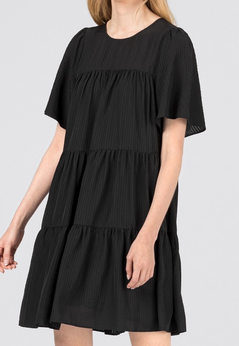 Robe noire à volants en tissu texturé, dotée de manches courtes, d'un col rond et d'une coupe ample. La longueur arrive au-dessus des genoux.