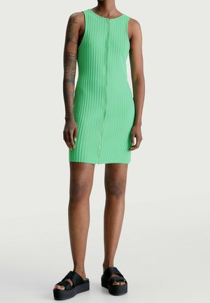 Persona con un vestido mini verde sin mangas y acanalado, con cremallera frontal y sandalias negras de plataforma, de pie frente a un fondo liso.