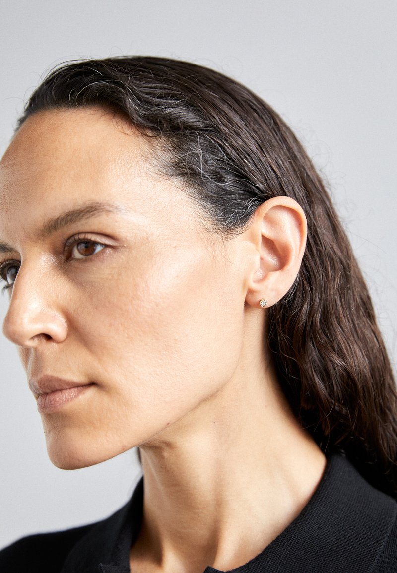 Kolczyk w kształcie okręgu, wyłożony diamentami, wykonany z metalu srebrnego, gładka faktura, zaprezentowany na fakturowanej czarnej odzieży na neutralnym tle.