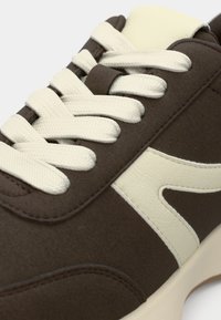 Καφέ αθλητικό παπούτσι με κρέμ κορδόνια, διαθέτοντας λεία υφή, λογότυπο σε κρέμ χρώμα και απαλή στρογγυλεμένη σχεδίαση στη μύτη.