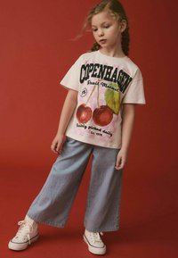 Hvid t-shirt med et kirsebærmotiv og teksten "Copenhagen Fruit Market"; parret med blå stribede vide bukser; hvide sneakers.