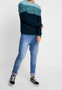 Stickad tröja i teal och mörkblå med strukturerat mönster; kombinerad med ljusblå jeans och svarta sneakers med vita accenter.