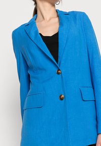 Modrý lněný blazer se zapínáním na jedno tlačítko, výstřihovými klopami a dvěma předními kapsami. Hladká textura s uvolněným střihem.