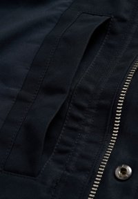 Mornarsko modra tkanina z gladko teksturo ima zadrgo na odprtini in stranski žep. Šivi so izraziti ob robovih za vzdržljivost.